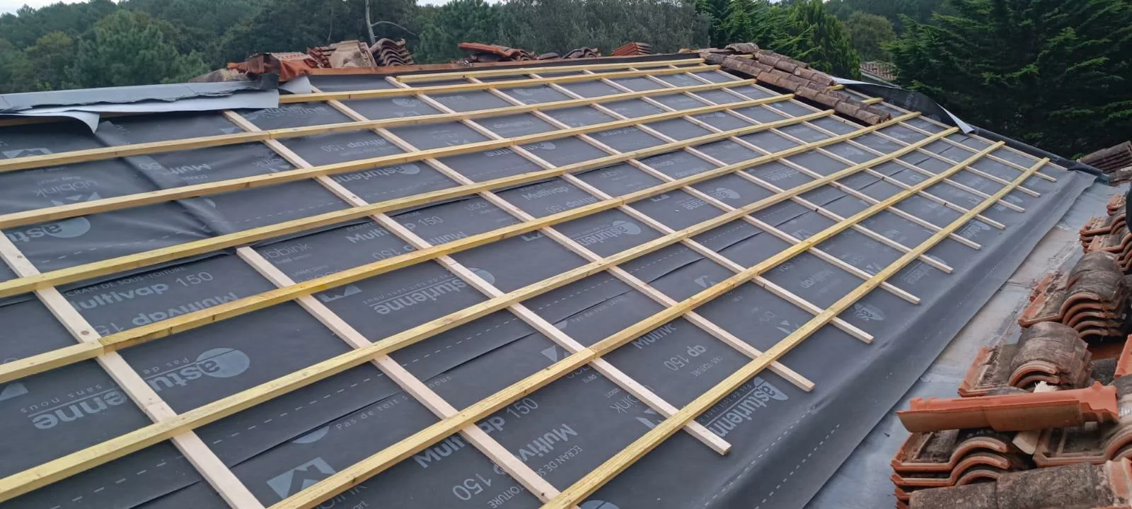 Toiture en cours de rénovation avec la pose d’un écran sous-toiture respirant Multivap 150 et l’installation d’un litelage en bois destiné à accueillir des tuiles canal, sur une charpente inclinée dans un environnement forestier.