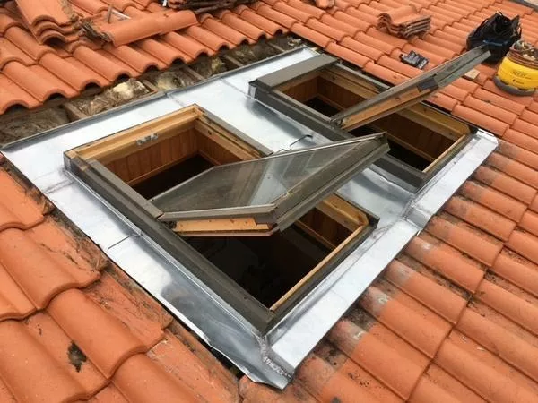 En premier plan, deux fenêtres de toit à ouverture battante, intégrées dans une couverture en tuiles canal rouges traditionnelles. Les encadrements en bois sont visibles, avec un habillage en zinc assurant l’étanchéité autour des châssis. En second plan, on distingue les tuiles partiellement déposées, indiquant un chantier de rénovation de toiture. Exemple de pose de fenêtres de toit avec raccords zinc sur toiture ancienne en tuiles terre cuite.